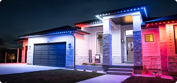 Modern house at night with red, white, and blue LED lights outlining the roof, a dark garage door, and a "Welcome" sign by the front entrance.
