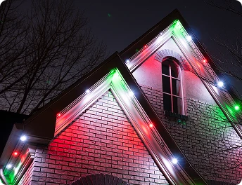 The peaked roof of a brick house is decorated with red, green, and white lights at night; bare tree branches are visible in the background.