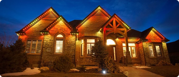 A single-story brick house is illuminated by red, yellow, and green lights along the roofline at dusk.