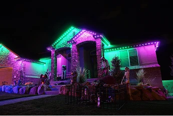 A single-story house is decorated with purple and green lights at night, featuring Halloween props like skeletons and gravestones in the front yard.
