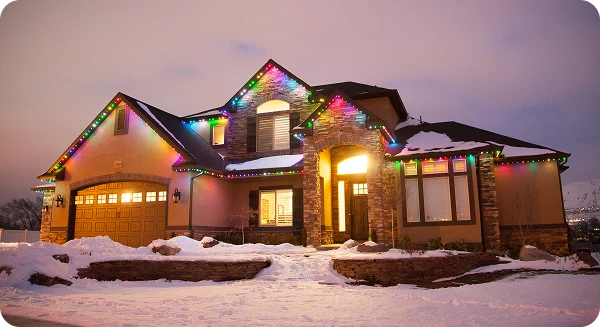 A two-story house with snow on the ground and colorful holiday lights decorating the roofline, illuminated at dusk.