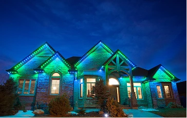 A brick house at night is illuminated by green and blue LED lights along the roofline and architectural features.