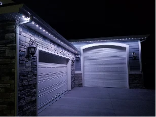 Two closed garage doors on a house exterior at night, illuminated by white LED lights along the roofline and wall-mounted lanterns.