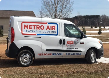 A white Metro Air Heating & Cooling service van is parked on a dirt driveway in front of a house with lawn and trees in the background.